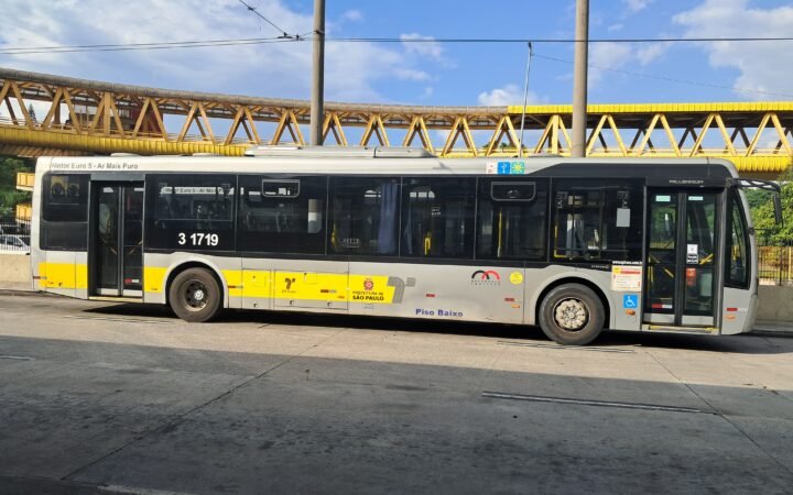Ônibus municipal amarelo e cinza da SPTrans, da empresa Metrópole Paulista, parado em uma via de São Paulo sob uma passarela metálica