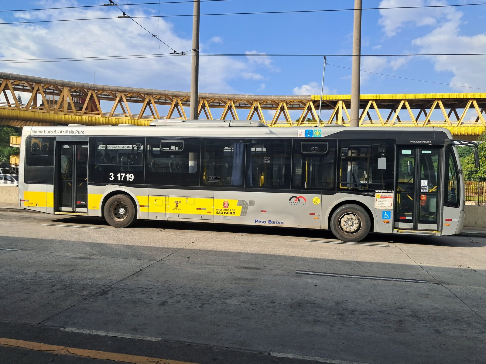 Ônibus municipal amarelo e cinza da SPTrans, da empresa Metrópole Paulista, parado em uma via de São Paulo sob uma passarela metálica