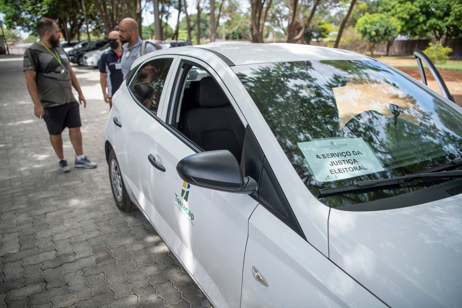 Carro branco com aviso no para-brisa 'A serviço da Justiça Eleitoral' estacionado em pátio arborizado, com três homens ao fundo.
