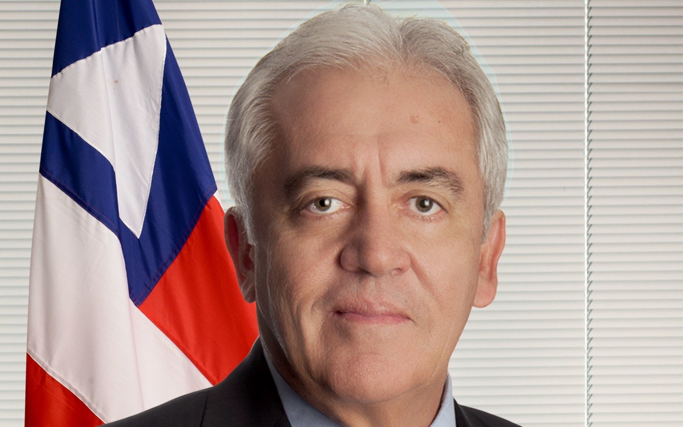 Retrato do senador Otto Alencar, de cabelos grisalhos, terno escuro e gravata listrada, diante da bandeira da Bahia.
