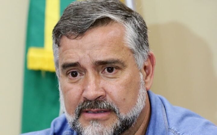 Retrato do deputado Paulo Pimenta, de barba grisalha e camisa azul, diante de uma bandeira do Brasil