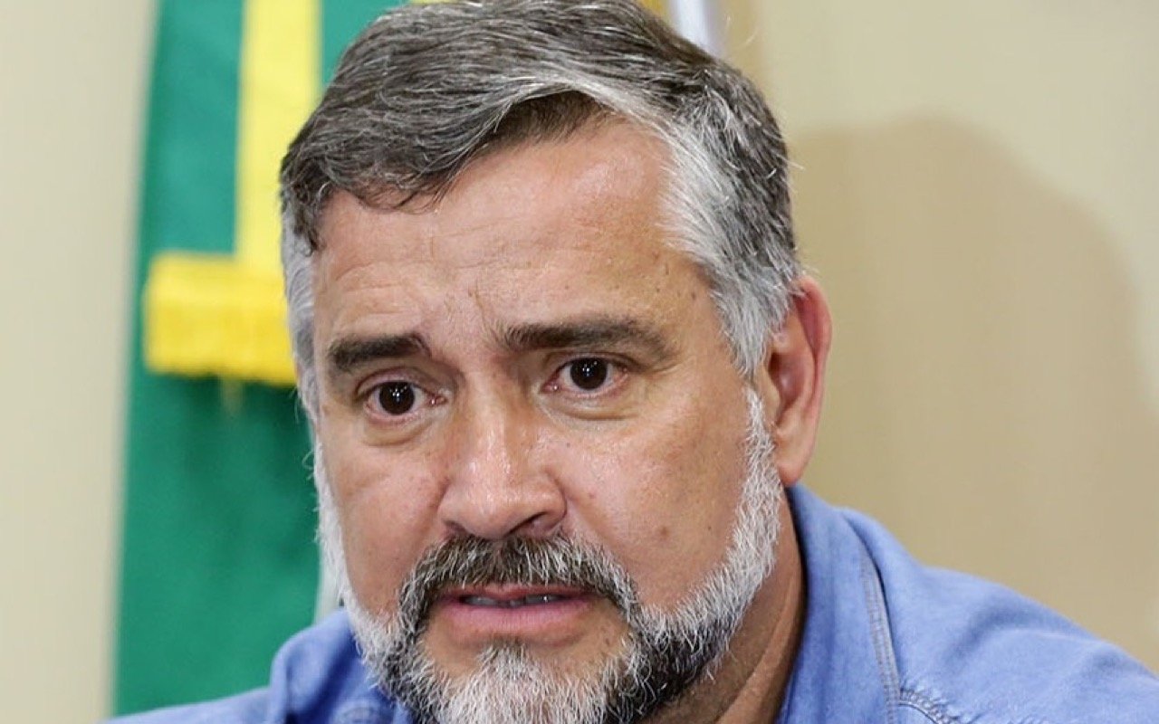 Retrato do deputado Paulo Pimenta, de barba grisalha e camisa azul, diante de uma bandeira do Brasil