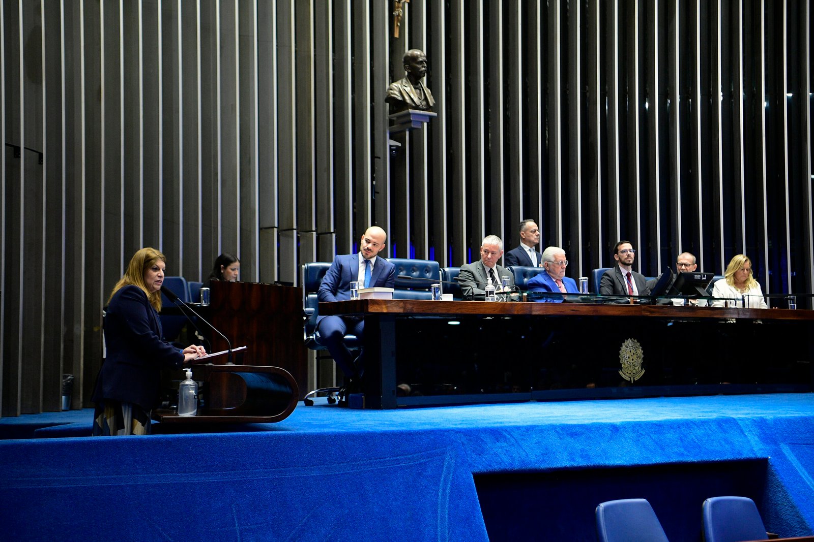Plenário do Senado Federal com a mesa diretora ao fundo e uma oradora na tribuna, sob painel de madeira ripada