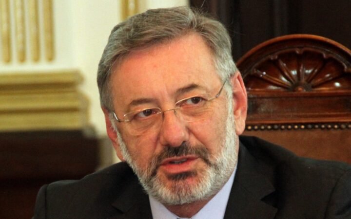 Retrato de Márcio Elias Rosa, de óculos e barba grisalha, falando durante uma reunião