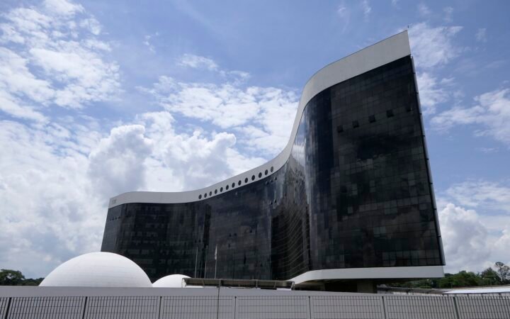 Edifício-sede do Tribunal Superior Eleitoral, de fachada curva em vidro escuro, sob céu azul com nuvens, em Brasília
