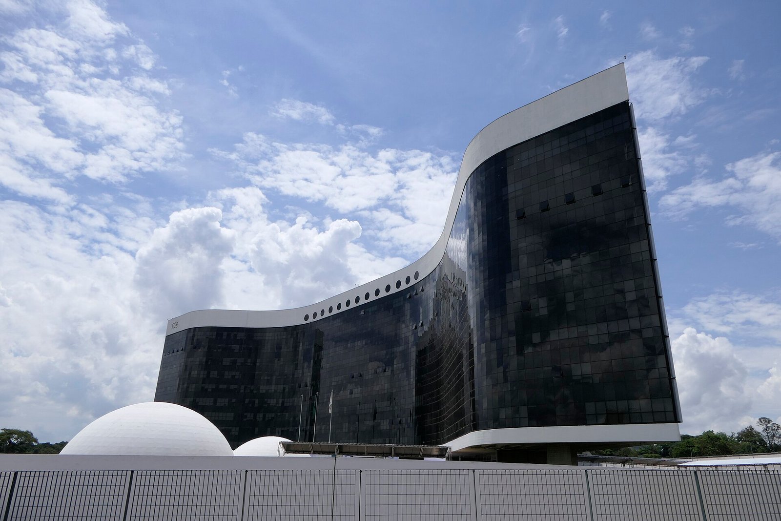 Edifício-sede do Tribunal Superior Eleitoral, de fachada curva em vidro escuro, sob céu azul com nuvens, em Brasília