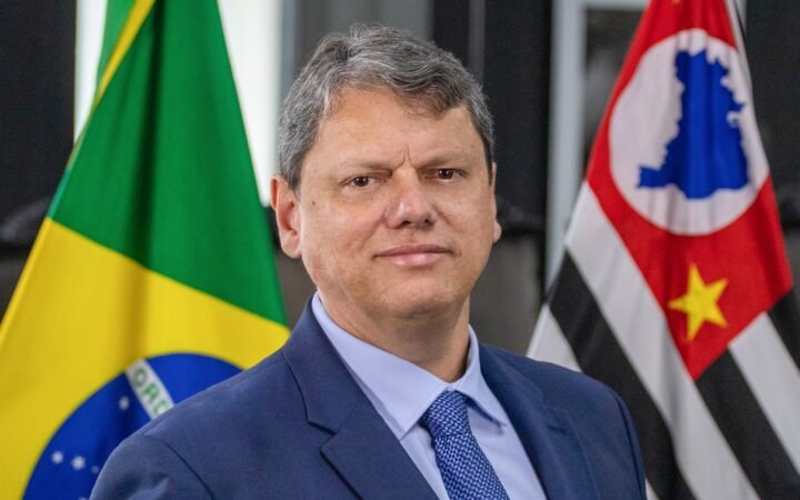 Retrato do governador de São Paulo, Tarcísio de Freitas, de terno azul, entre as bandeiras do Brasil e do estado de São Paulo