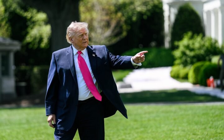O presidente dos EUA, Donald Trump, de terno azul e gravata rosa, aponta com a mão direita num gramado da Casa Branca.
