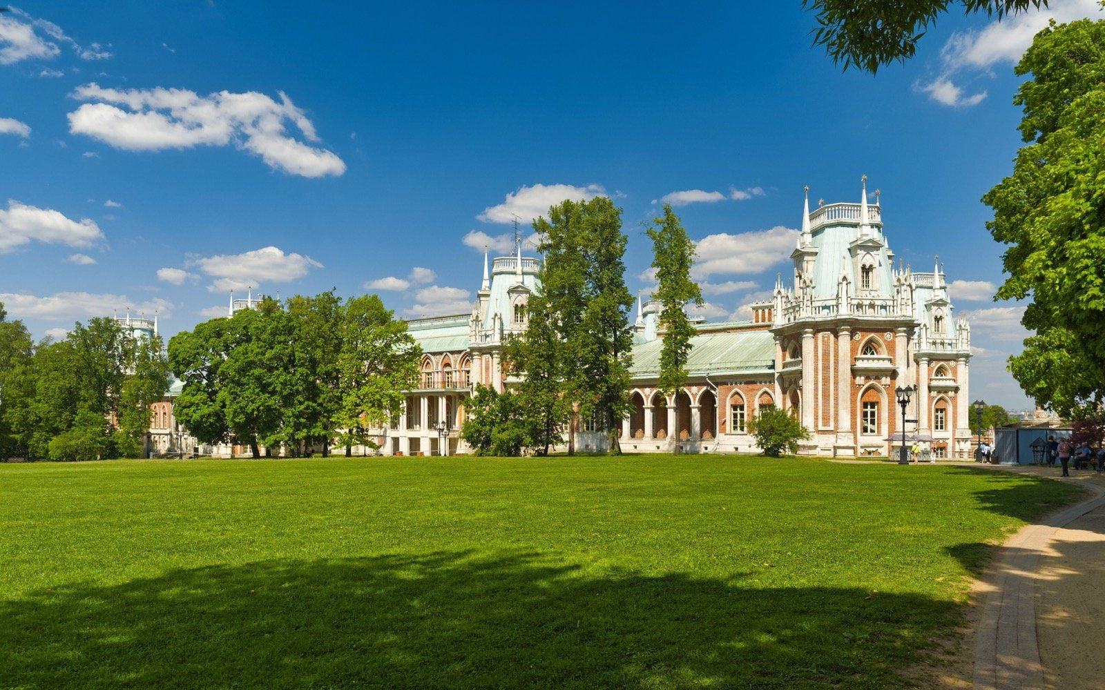Fachada em tijolo vermelho e pedra branca do Grande Palácio de Tsaritsyno, em Moscou, com gramado e árvores à frente sob céu azul.