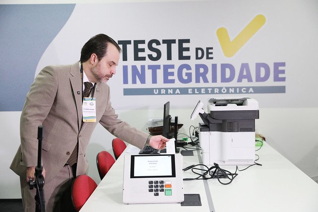 Servidor da Justiça Eleitoral opera urna eletrônica durante o teste de integridade no TSE, diante de painel com os dizeres Teste de integridade