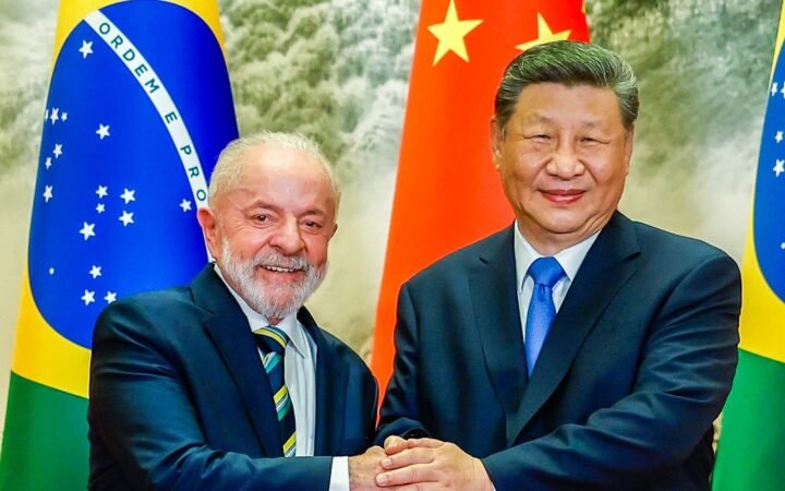 Lula e Xi Jinping, sorrindo, de mãos dadas diante de bandeiras do Brasil e da China.